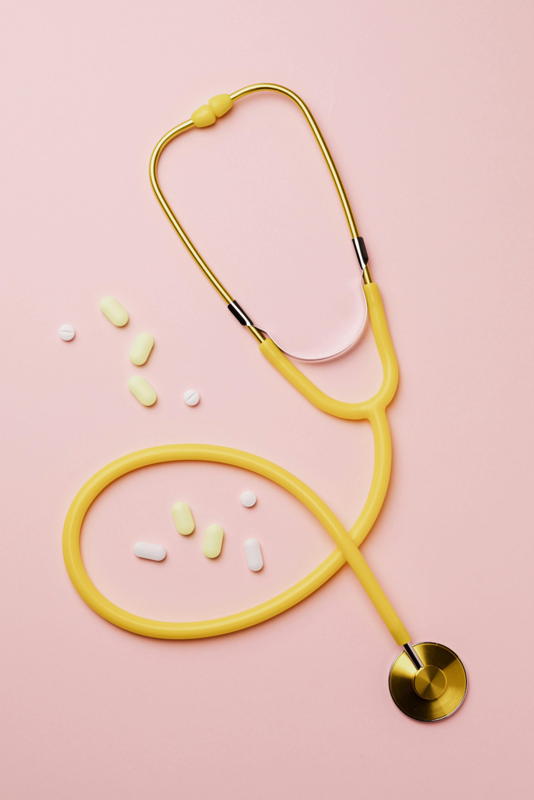 Flat lay of a yellow stethoscope and assorted pills on a pastel pink background.